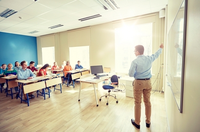 Das Bild zeigt einen lichtdurchfluteten Klassenraum. Der Lehrer im Vordergrund steht vor der Klasse und erklärt etwas.