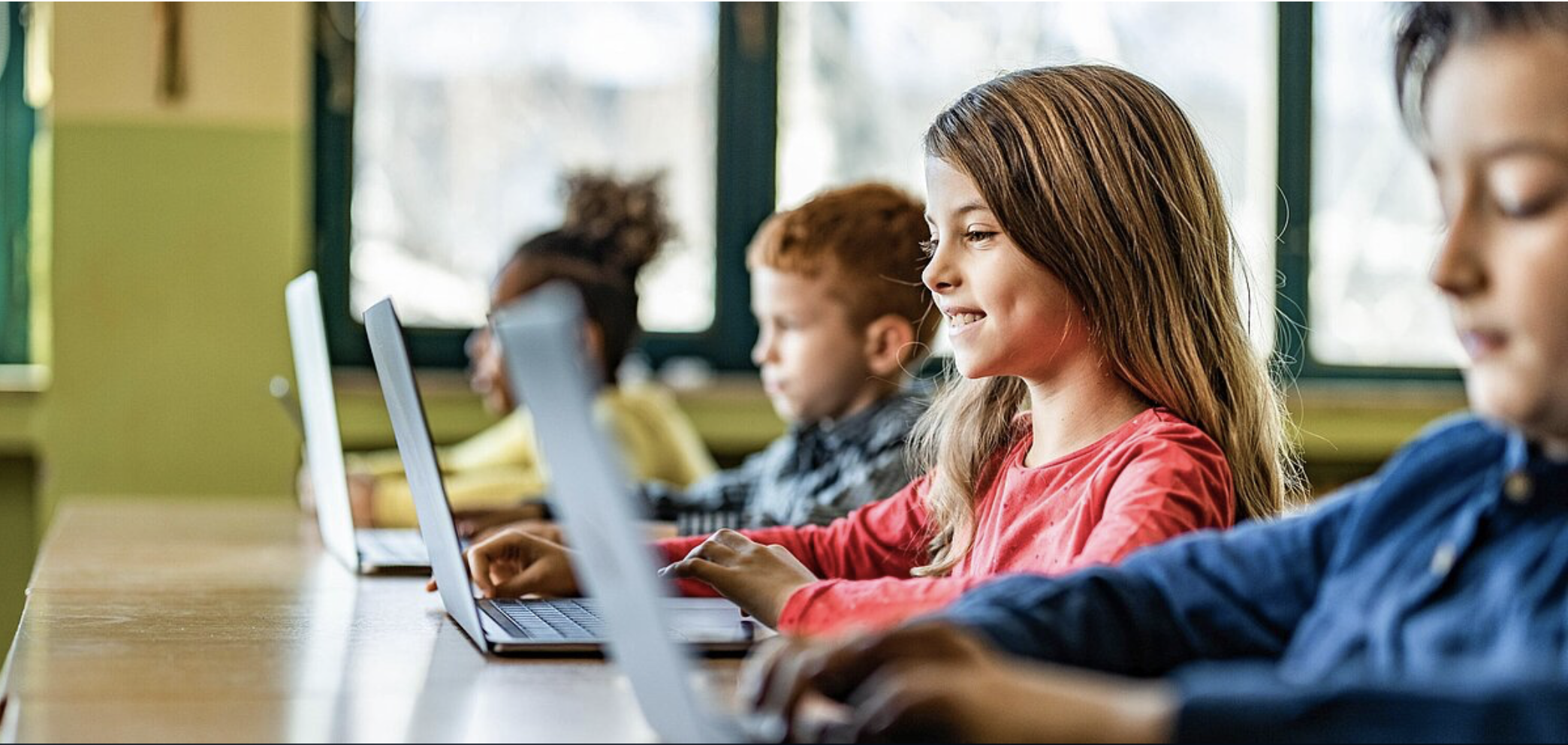 Eine Gruppe von Grundschulkindern sitzt nebeneinander in einem Klassenzimmer und arbeitet an Laptops. Ein Mädchen mit blonden Haaren in einem roten Pullover lächelt, während sie tippt. Im Hintergrund sitzen weitere Kinder an ihren Geräten.