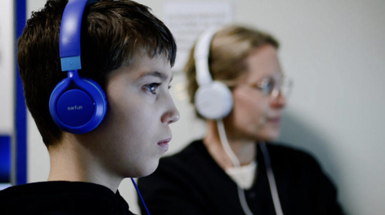 Ein Schüler mit blauen Kopfhörern arbeitet konzentriert am Computer. Im Hintergrund sitzt eine Lehrkraft mit weißen Kopfhörern