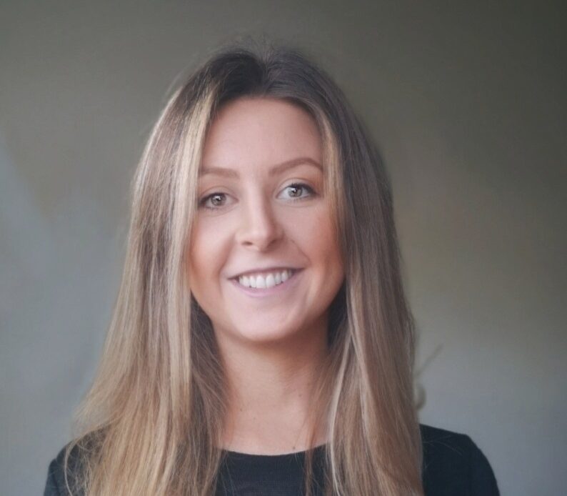 Dr.in Sally Kröger trägt ein schwazes langärmeliges Oberteil und hat lange braun/blonde Haare, die sie offen trägt. Sie schaut mit einem offenen breitem Lächeln in die Kamera.