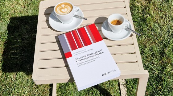 Der Sammelband "Transfer in Pädagogik und Erziehungswissenschaft II" liegt auf einem Tisch in der Sonne, daneben stehen zwei Tassen Kaffee; im Hintegrund ist grüner Rasen zu sehen.