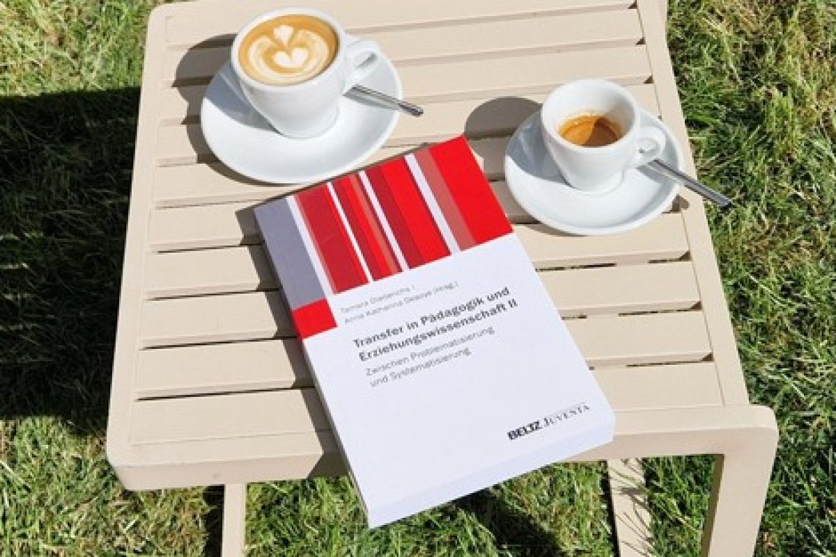 Der Sammelband "Transfer in Pädagogik und Erziehungswissenschaft II" liegt auf einem Tisch in der Sonne, daneben stehen zwei Tassen Kaffee; im Hintegrund ist grüner Rasen zu sehen.